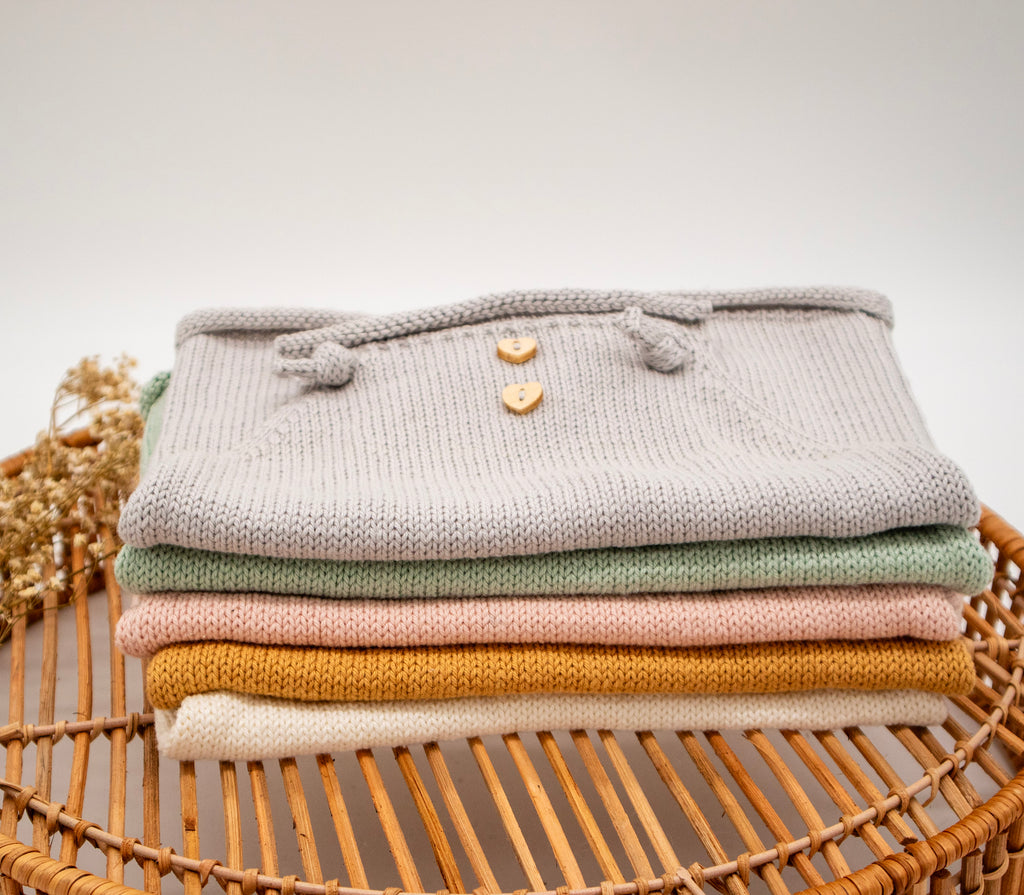 Gestapelte Vintage-Handgestrickter-Neugeborenen-Strampler in Grau, Safran, Rosa und Mint, präsentiert auf einem Bambuskorb.