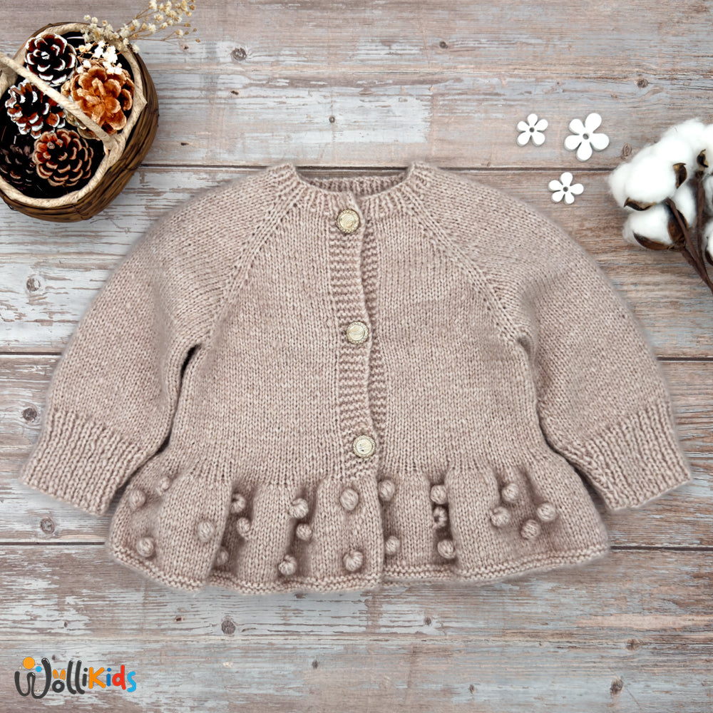 Handgestrickte cremefarbene Baby-Strickjacke mit Popcorn-Details und drei goldenen Knöpfen, auf rustikalem Holzhintergrund präsentiert.