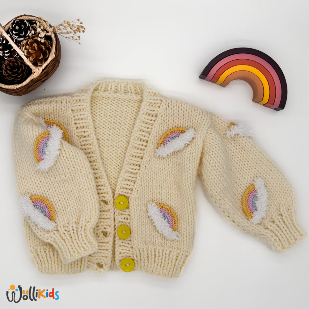 Handgestrickte Regenbogen-Strickjacke in Creme mit gelben Knöpfen und Regenbogen-Design.