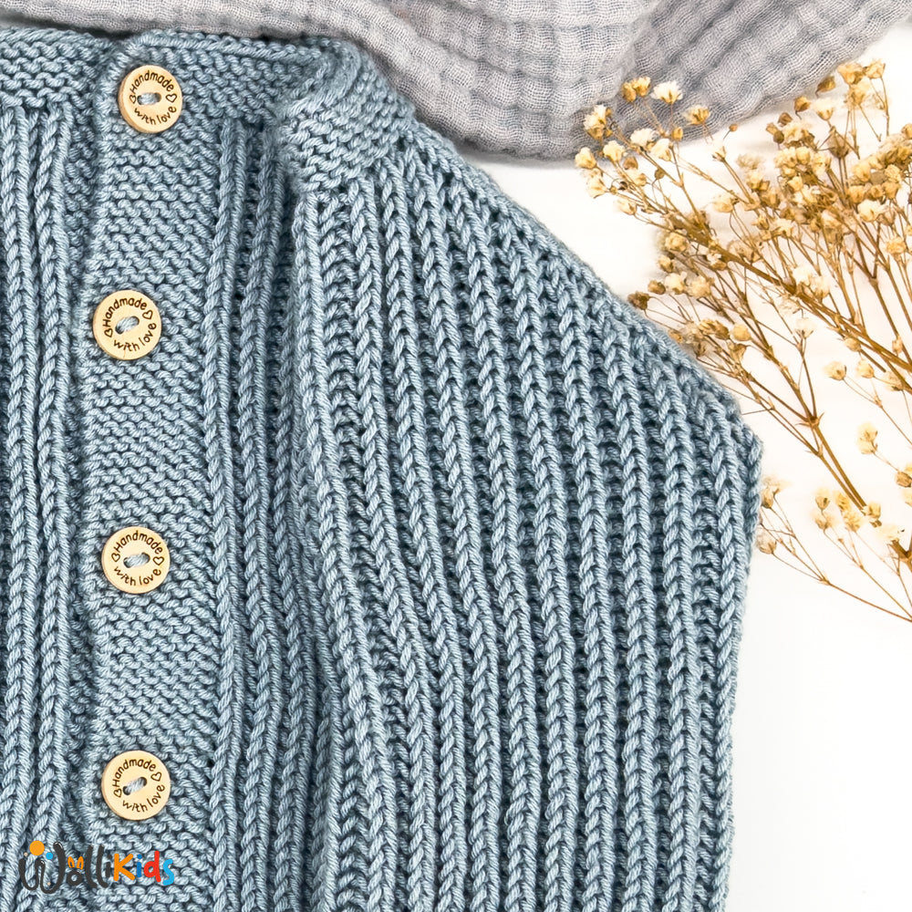 Detaillierte Nahaufnahme der blauen Vollpatent Babystrickjacke mit handgearbeiteten Knöpfen und feiner Strickstruktur, dekoriert mit Trockenblumen und Wollikids-Logo.

